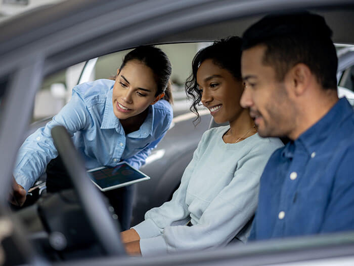 Two people sit inside a car while a salesperson shows them features